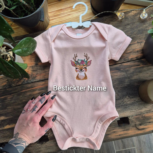 Bestickter Babybody rosa mit Reh Motiv liegt auf Holztisch neben Pflanzen